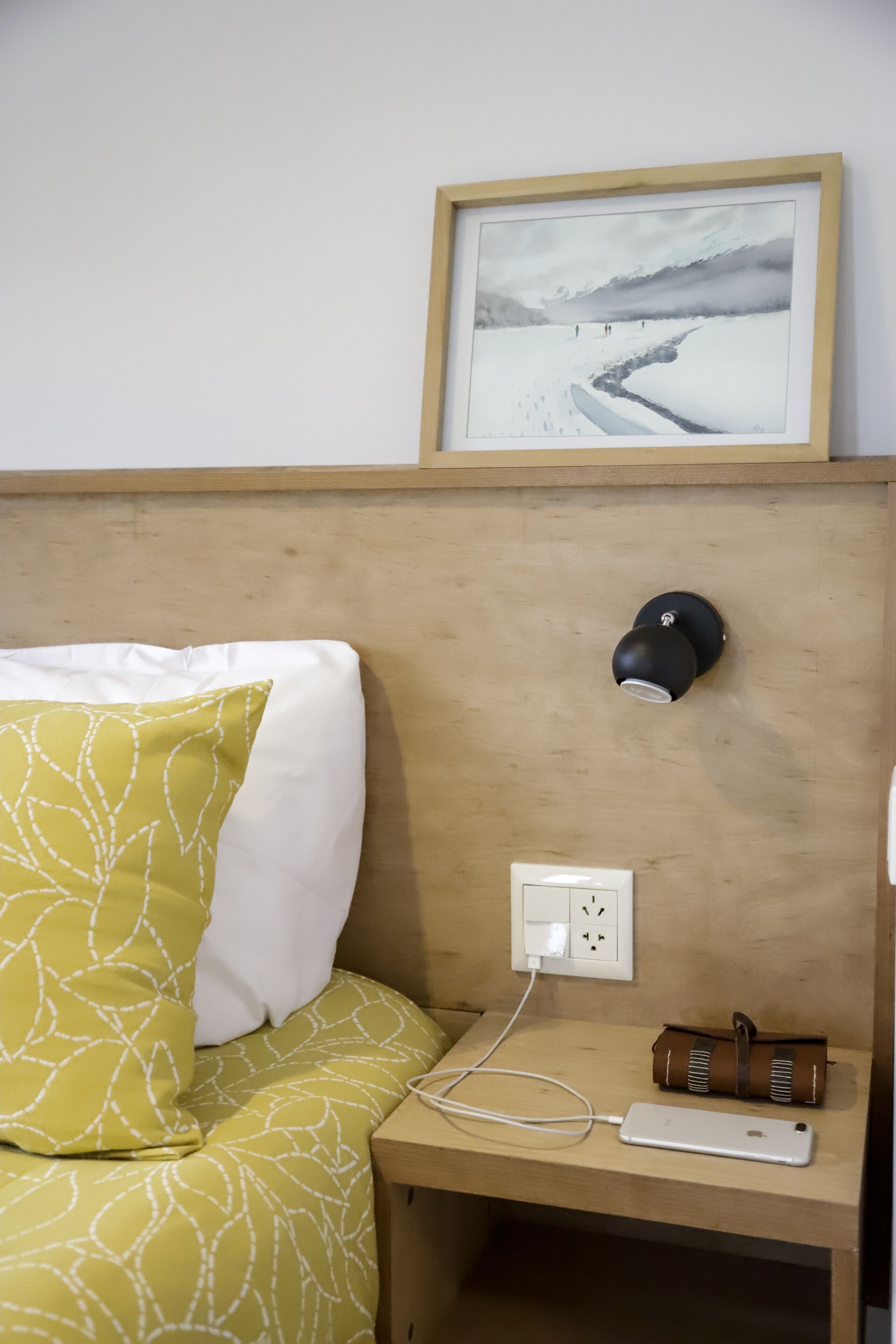 Een net opgemaakt bed met een kussen in geel en wit patroon, een nachtkastje met een stopcontact, een oplader voor de telefoon en een lijst aan de muur.