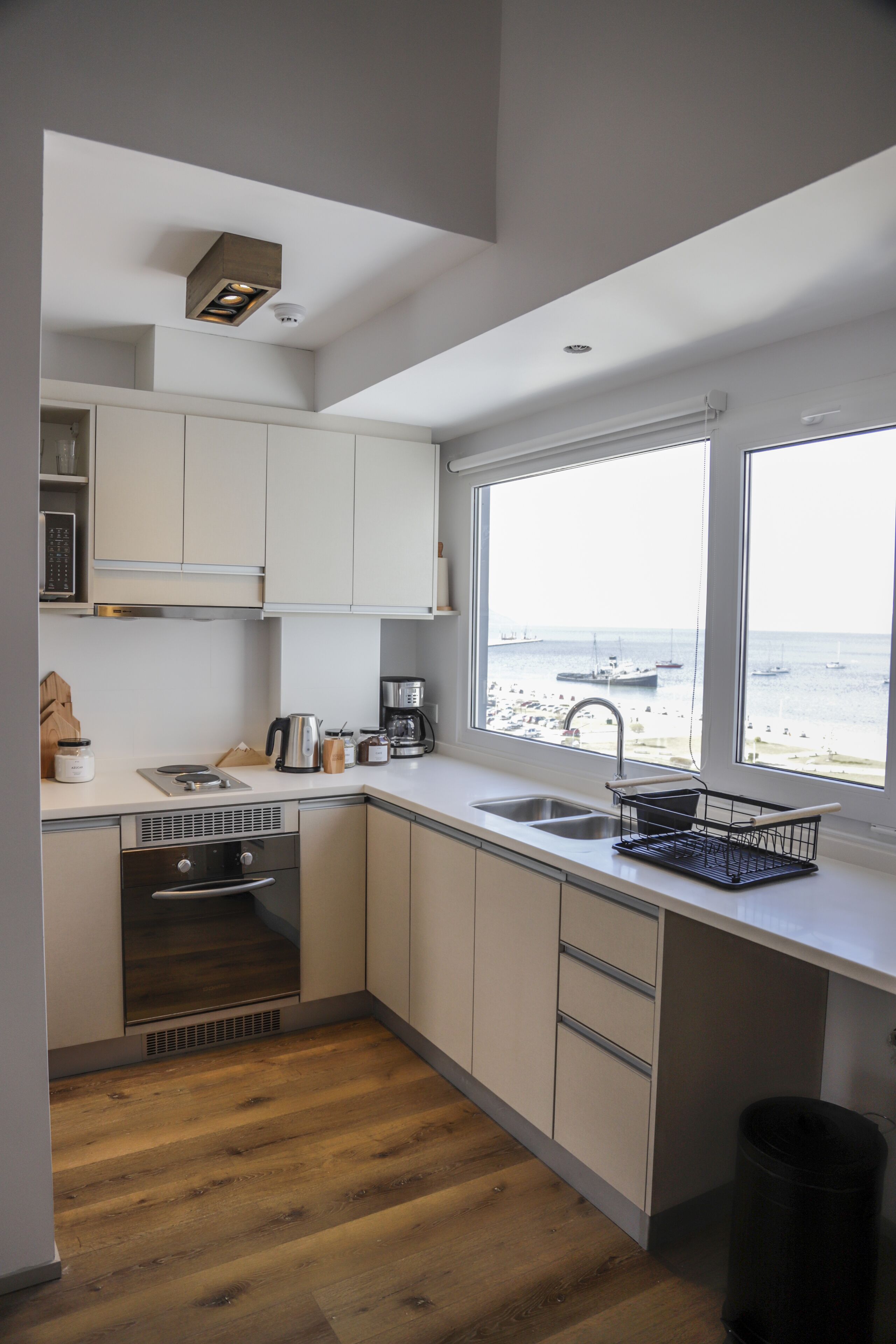 A modern kitchen with a view of the sea, featuring white cabinets, a sink, and a window overlooking the beach.
