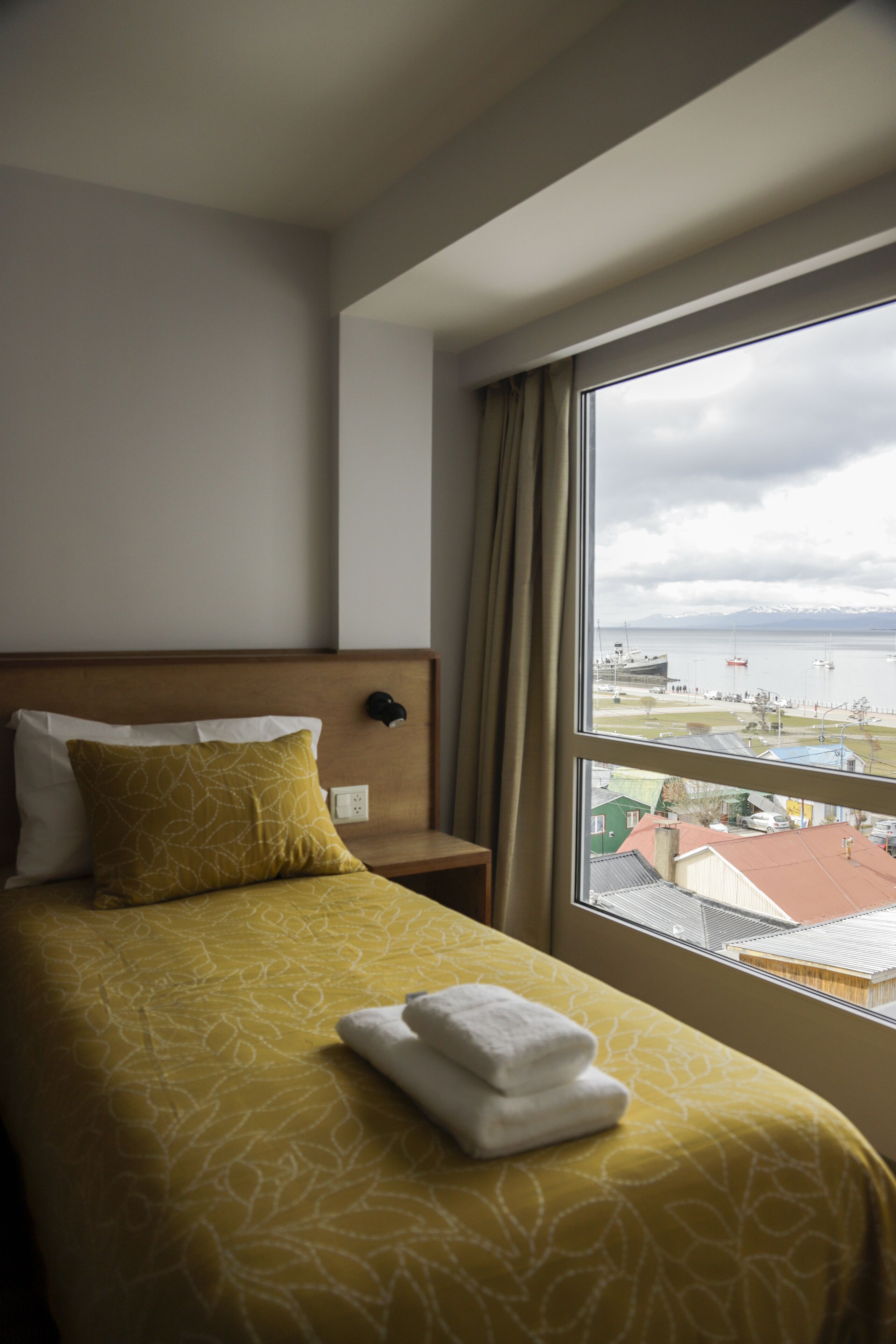 A hotel room with a bed, a nightstand, and a window with a view of a body of water and buildings.