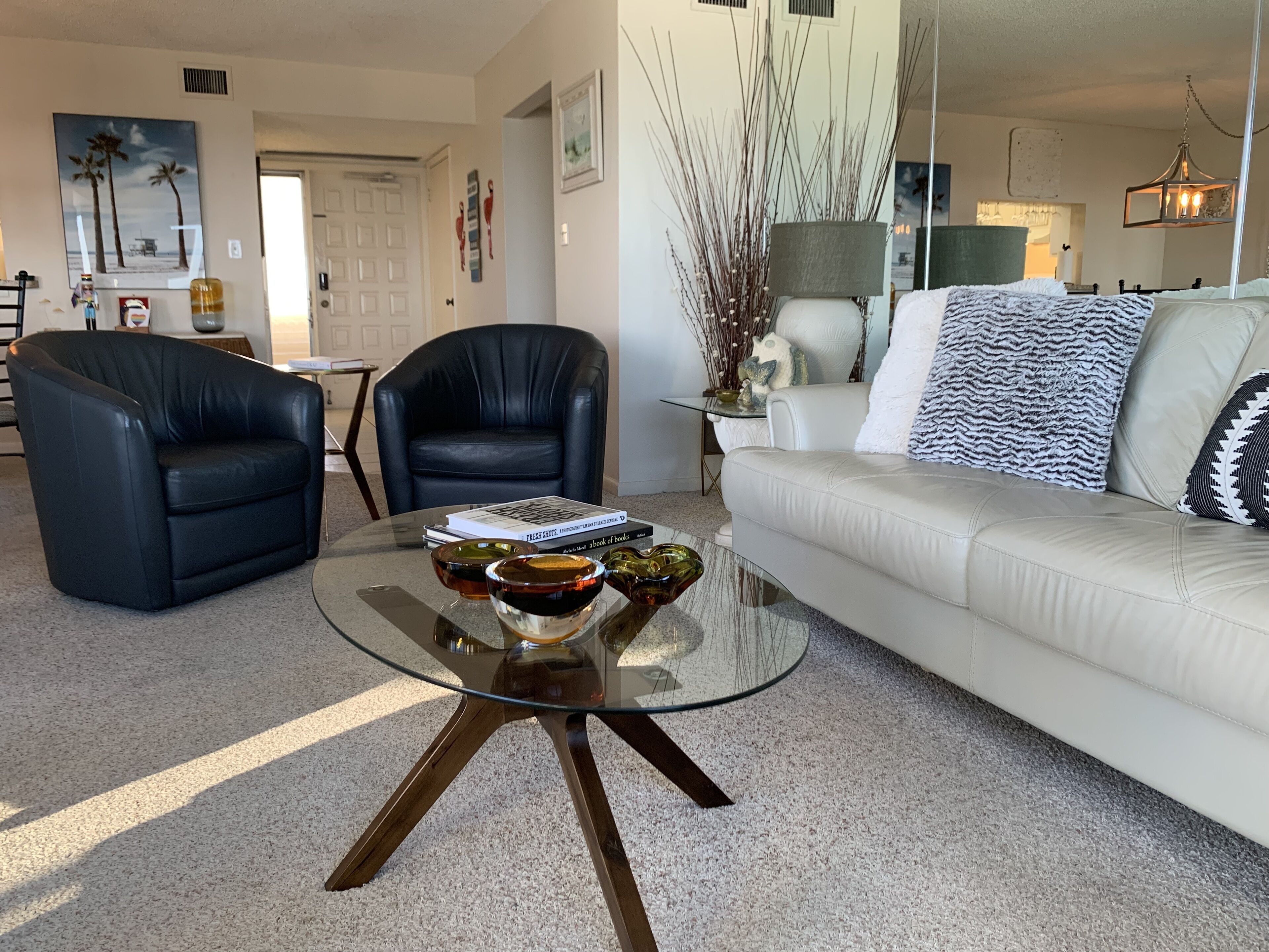 A living room with a glass coffee table, two leather armchairs, a white sofa, and a mirror reflecting the room.