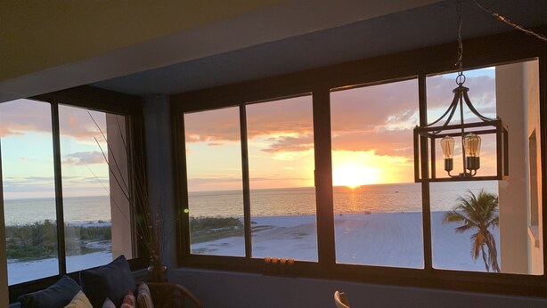 Una stanza con una grande finestra vista spiaggia al tramonto. C'è un divano in vimini con cuscini e un piccolo tavolino con una lampada.