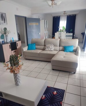 A modern living room with a beige sectional sofa, a white coffee table, and a vase with flowers. There is a cabinet with a plant, a door labeled "SALLE DE RECEPTION," and a dining area in the background. - Villa Saphira (Antananarivo)