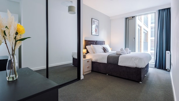 A modern hotel room with a large bed, bedside tables, a mirror, and a view of the city through the window. - 3 Bedroom Apartment (Manchester)