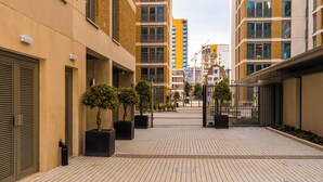 A modern urban courtyard with a paved walkway, potted trees, and contemporary buildings in the background.