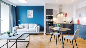 A modern living room with a sofa, dining table, and chairs, a kitchen with appliances, and a blue accent wall.