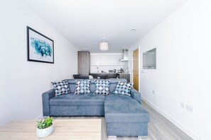 Uma sala de estar moderna com um sofá modular cinza, uma mesa de centro com uma planta e uma cozinha ao fundo.