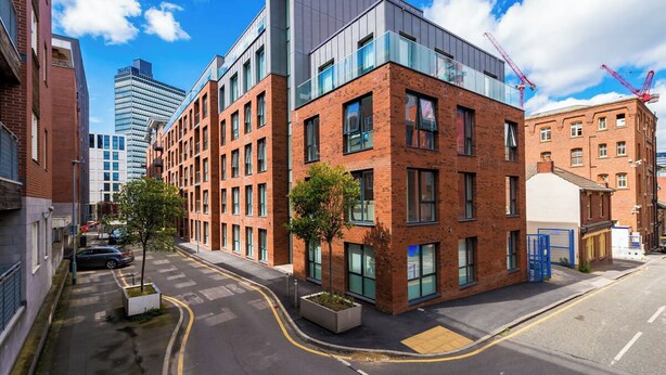 A modern urban street with a mix of commercial and residential buildings, including a prominent red-brick building with large windows and a flat roof.