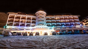 A hotel with a central tower, multiple balconies, and a parking lot with cars. - Sporthotel All-Inclusive (Silz)