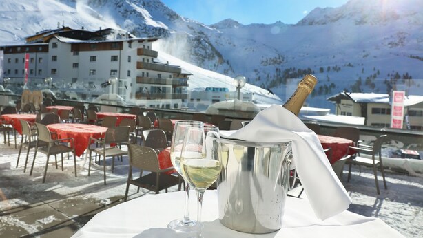A table with a bottle of champagne and a glass of white wine, set against a snowy mountain backdrop. - Sporthotel All-Inclusive (Silz)