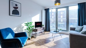 A modern living room with a blue armchair, a grey sofa, a coffee table, and a TV on a stand. - 2 Bedroom Apartment (Manchester)