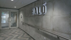 Un corridoio con un cartello metallico che riporta la scritta "HALO", un ingresso con porta in vetro e un pavimento a disegno a spina di pesce.