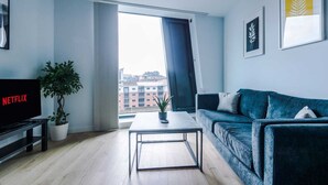 A modern living room with a sofa, a coffee table, a TV displaying 'NETFLIX', a potted plant, and a view of buildings through a large window. - 1 Bedroom Apartment (Manchester)