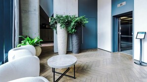 A modern lobby with a round table, white chairs, large planters with greenery, and a blue wall with an elevator. - 1 Bedroom Apartment (Manchester)