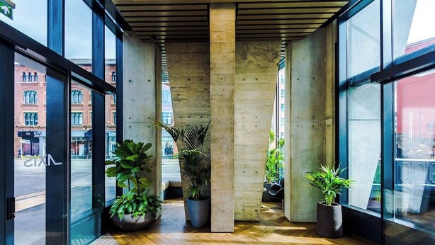A modern lobby with large glass windows, concrete pillars, and potted plants. - 1 Bedroom Apartment (Manchester)