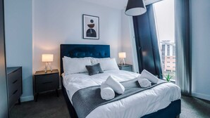 A modern hotel room with a large bed, a dark blue headboard, two bedside tables with lamps, a framed picture on the wall, and a view of the city through the window. - 1 Bedroom Apartment (Manchester)