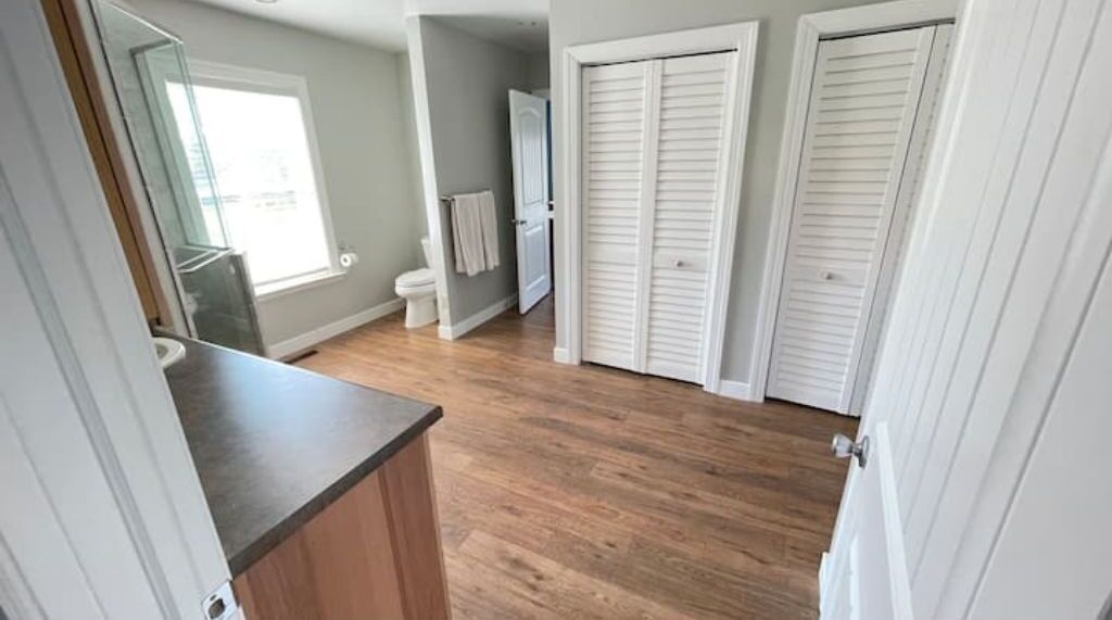 Une salle de bain avec un plancher en bois, un lavabo et un miroir. Il y a deux portes blanches et une serviette accrochée au mur.