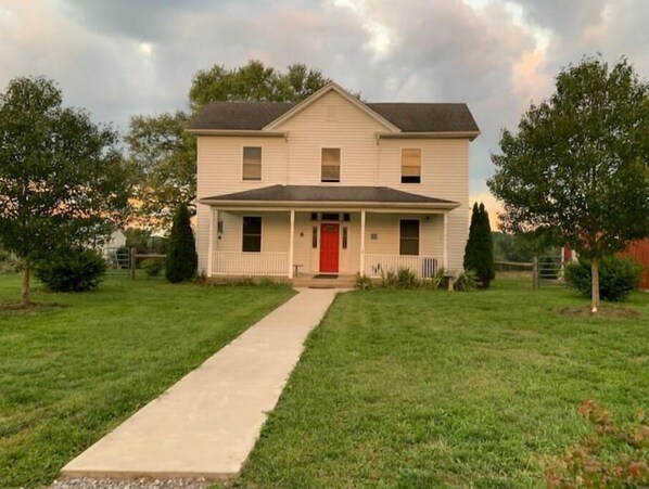 A two-story house with a red front door, a covered porch, and a front yard with a concrete path leading to the entrance. - Farmhouse at the foot of the Blue Ridge Mountains (Waynesboro)