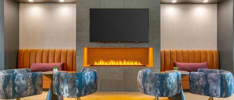 A modern lounge area with a flat-screen TV mounted above a fireplace, two sofas, and four unique blue chairs with gold bases.
