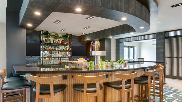 Een moderne bar met een gebogen plafond, houten barstoelen en een goed gevulde plank met diverse flessen.