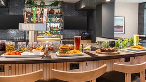 Een barruimte met diverse gerechten en dranken, waaronder hamburgers, friet en een hoog glas bier. Er zijn barstoelen en een bar met een biertappunt.