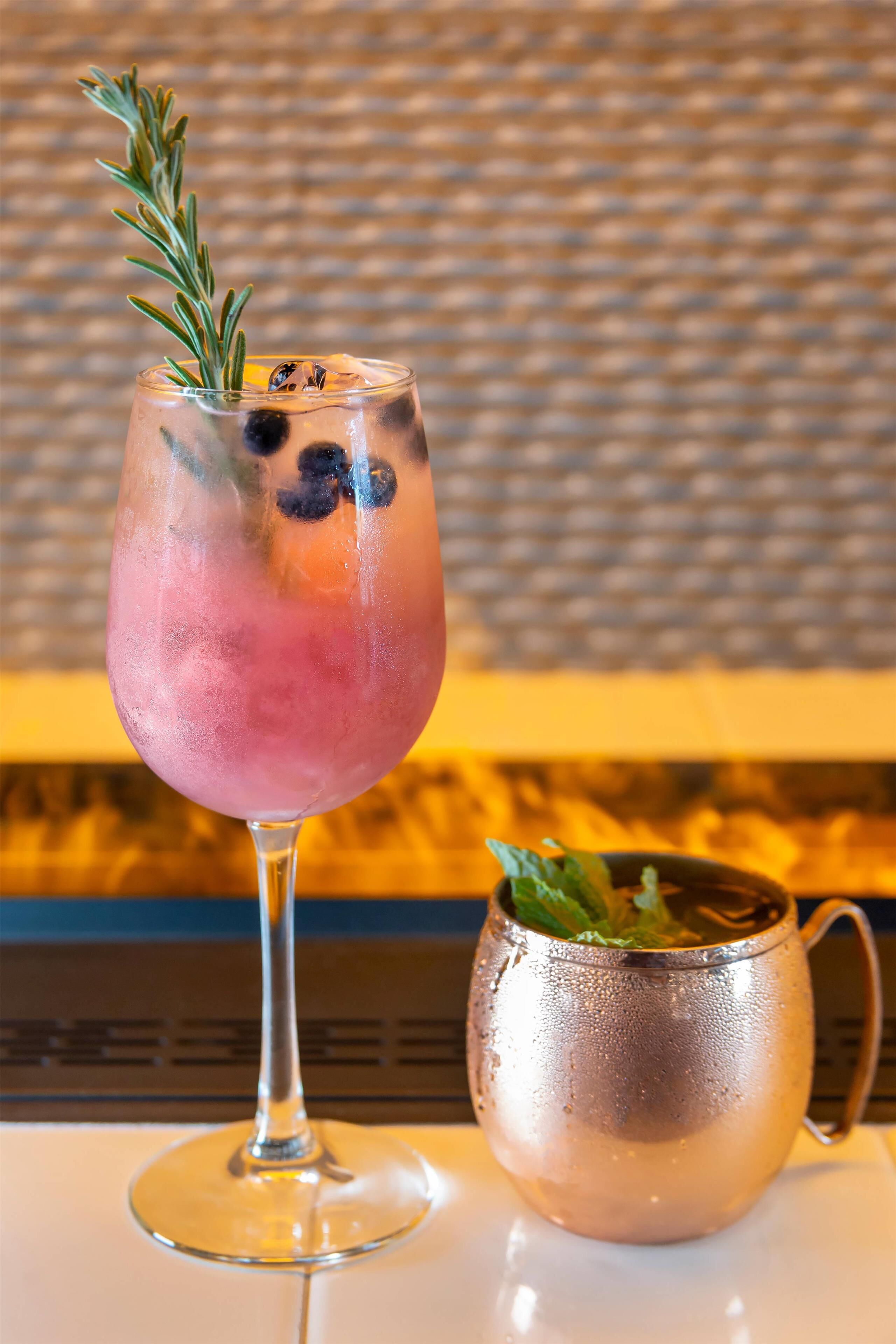 A cocktail glass with a sprig of rosemary and a blueberry garnish, next to a copper mug with mint leaves.