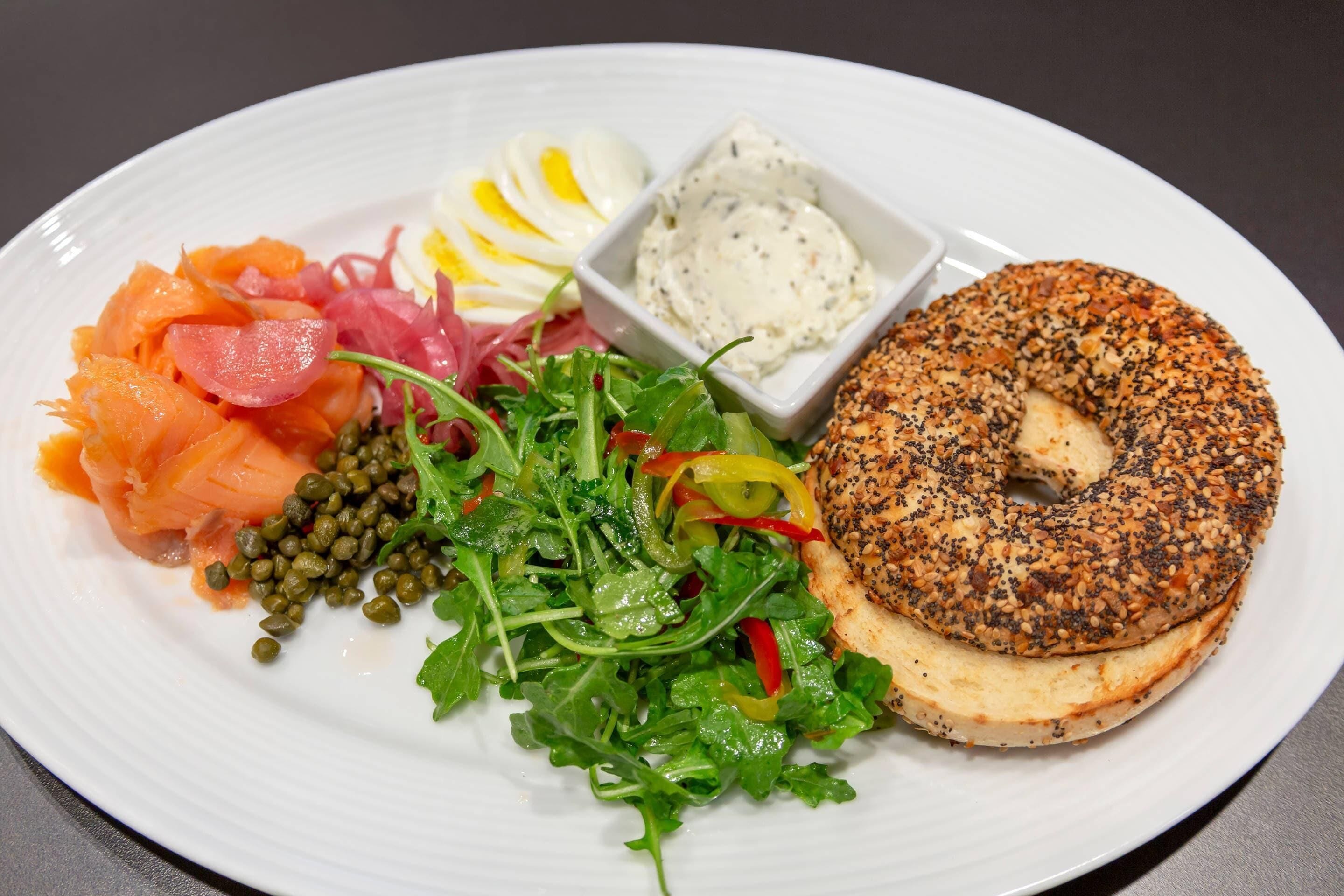 A plate with a bagel, smoked salmon, capers, a salad, hard-boiled eggs, and a small bowl of cream cheese.