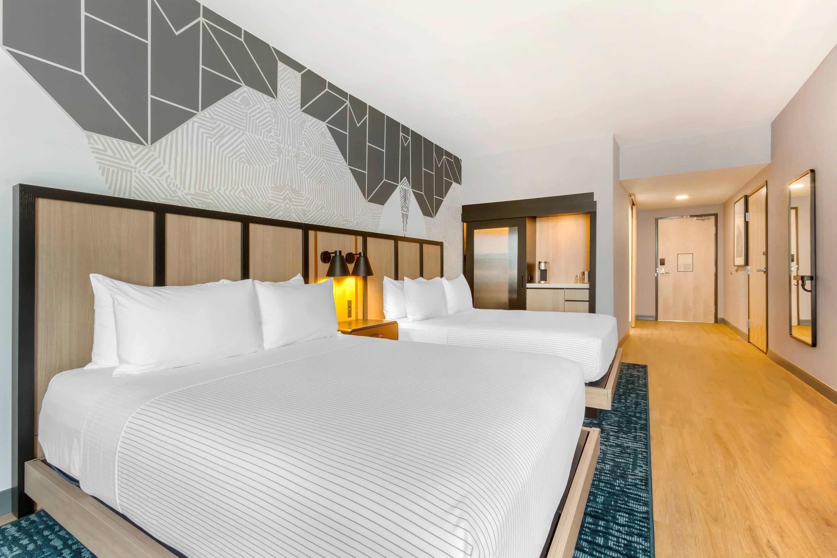 A hotel room with two beds, a large mirror, a wooden headboard, and a geometric wall design.