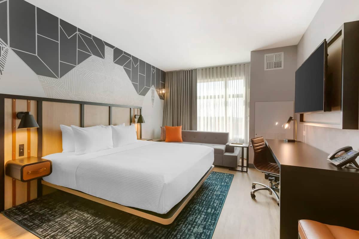 Ein Hotelzimmer mit einem großen Bett, einem Schreibtisch, einem Stuhl, einem Fernseher und einem Fenster mit Vorhängen.