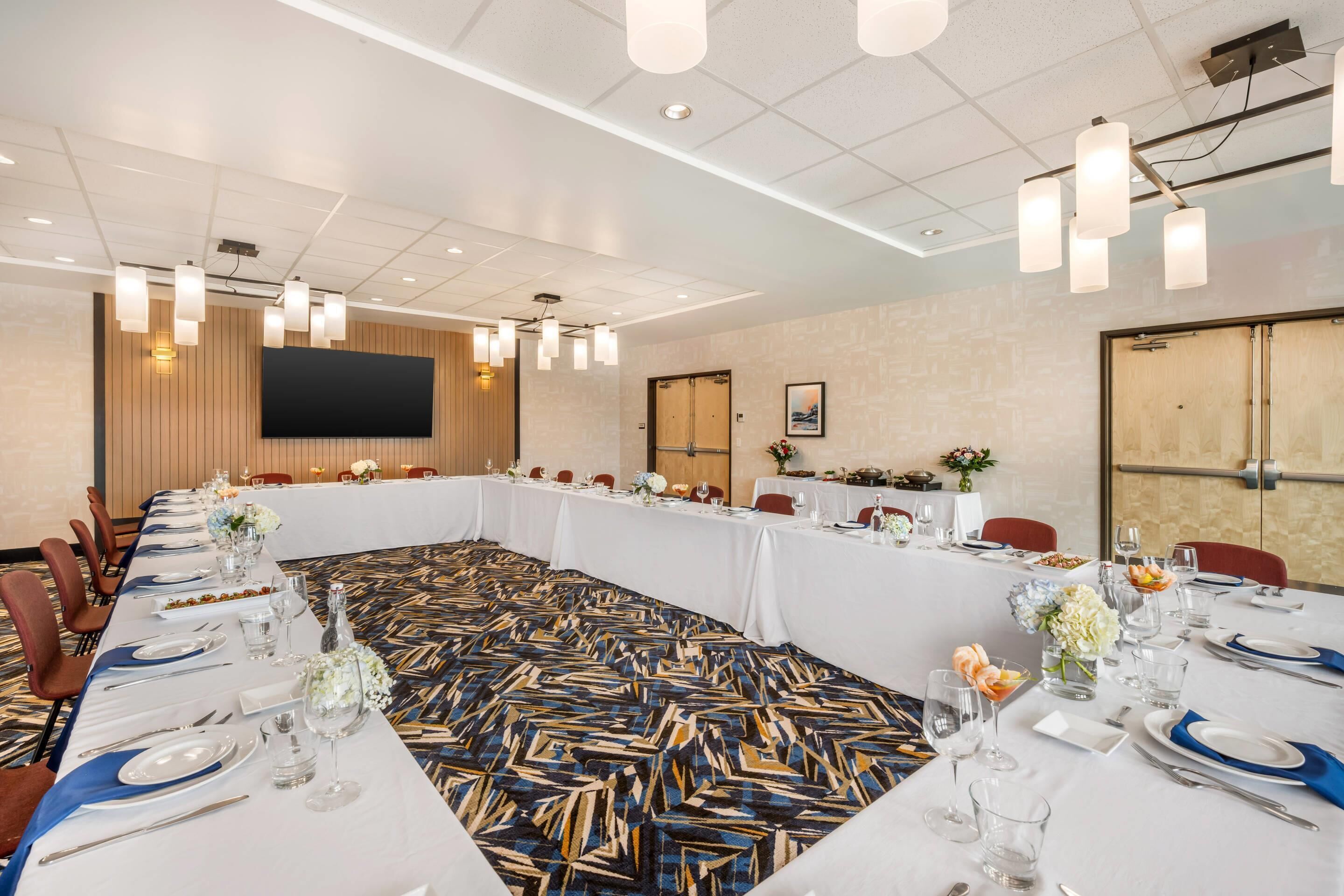 A conference room with a long table set for a meeting, equipped with chairs, a large screen, and modern lighting fixtures.