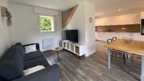 Ein modernes Wohnzimmer mit einem dunklen Sofa, einem kleinen runden Tisch und einem Fernseher auf einem weißen Schrank. Man sieht einen Essbereich mit einem Holztisch und Stühlen. Die Küche verfügt über weiße Schränke und eine Holz Arbeitsplatte.