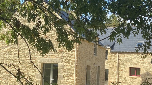 Ein Steinhaus mit einem großen Fenster und einer kleineren Tür, umgeben von Grünpflanzen.