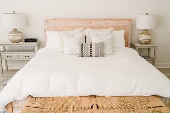 A neatly made bed with white bedding, a woven bench at the foot of the bed, and two bedside tables with lamps and a clock on one.