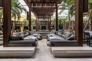 An outdoor seating area with sofas and tables, surrounded by palm trees and a pool.