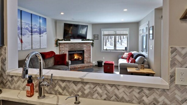 A modern living room with a fireplace, a large flat-screen TV, a sofa, and a kitchen area with a herringbone tile backsplash. - Ski Access from this Kettlebrook Condo with Modern Kitchen (Ludlow)