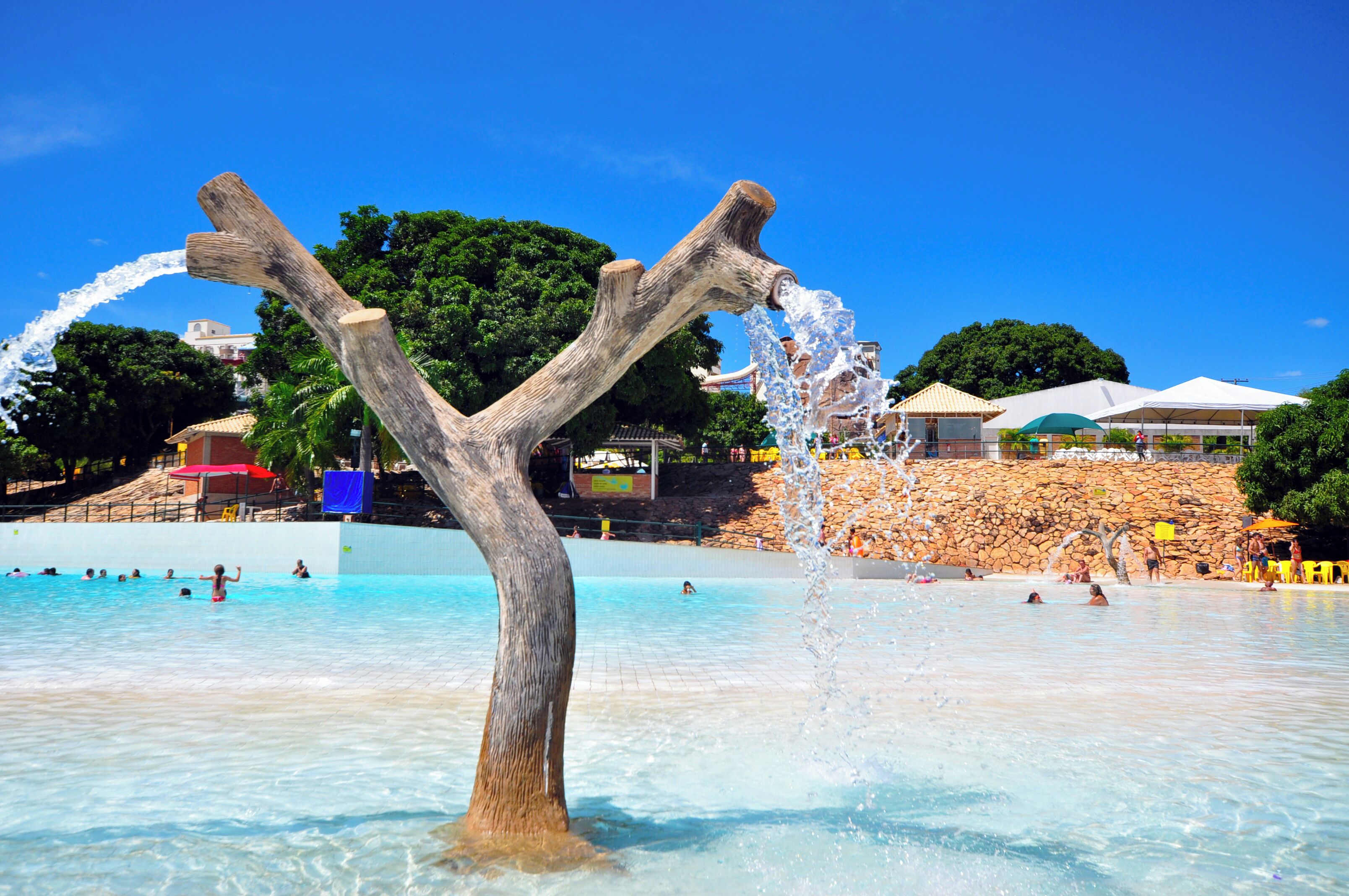 Ein Wasserspiel mit einer Holzkonstruktion in einem Poolbereich, umgeben von Menschen und einer Mauer mit Sonnenschirmen.