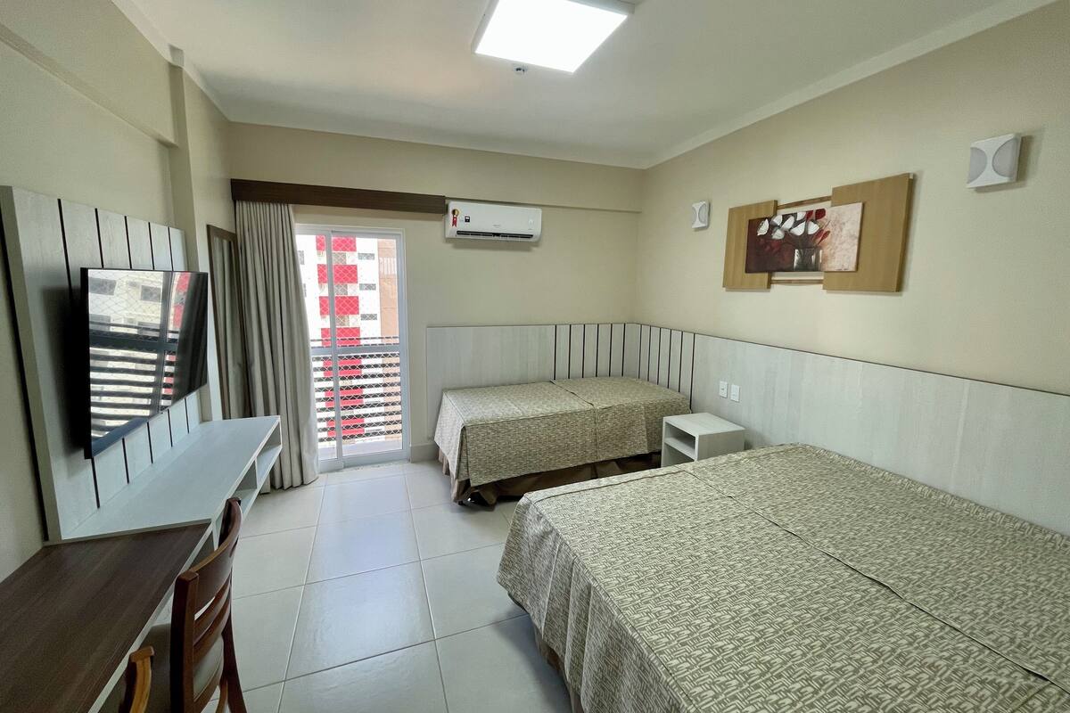 A hotel room with two beds, a desk, a chair, a TV, an air conditioner, and a window with a view of buildings.