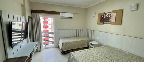 A hotel room with two beds, a desk, a chair, a TV, an air conditioner, and a window with a view of buildings.