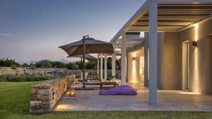 A modern outdoor patio area with a stone wall, lounge chairs, and a pergola with a large umbrella. - Leucosia - Favignana (Favignana)