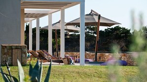 A covered outdoor seating area with lounge chairs and umbrellas, a stone wall, and a pool in the background.