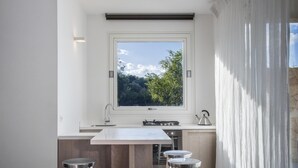 A modern kitchen with a large window, a central island with stools, and a view of greenery outside.
