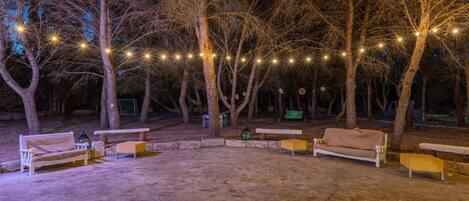 An outdoor seating area with benches and tables, surrounded by trees and string lights.