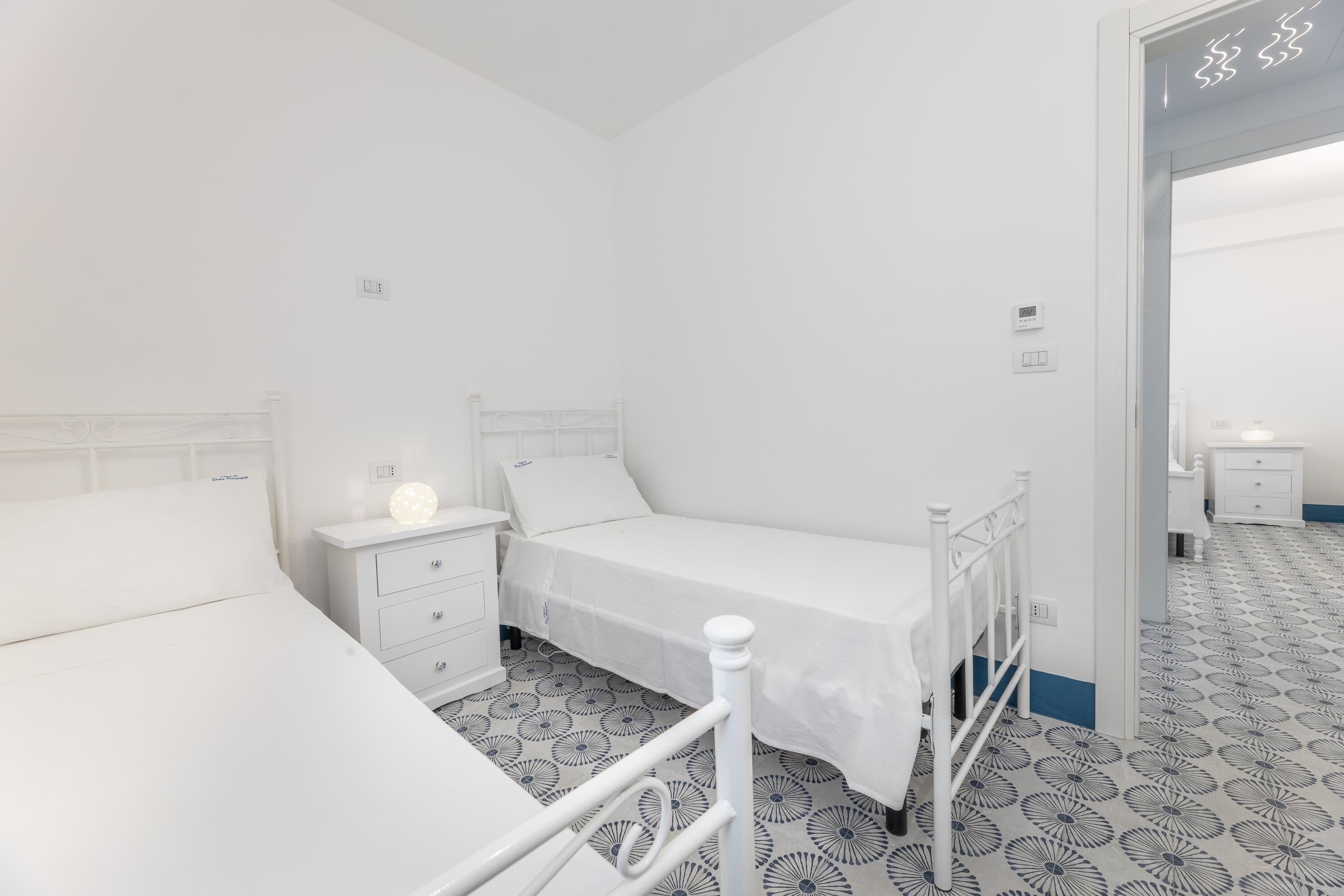 Ein kleines, weißes Zimmer mit einem Einzelbett, einem gemusterten Boden, einem Nachttisch und einem Beistelltisch.