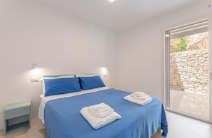 Ein Schlafzimmer mit einem großen Bett, blauer Bettwäsche, zwei weißen Handtüchern, einem Nachttisch und Blick auf eine Steinmauer durch eine Glastür.
