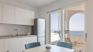 Een moderne keuken met witte kastjes, een gootsteen en een koelkast. Er staat een eettafel met stoelen en een decoratieve schaal erop. Een schuifpui leidt naar een balkon met uitzicht op zee.
