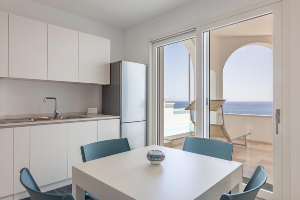 Una cocina moderna con armarios blancos, un fregadero y un refrigerador. Hay una mesa de comedor con sillas y un cuenco decorativo encima. Una puerta corrediza de vidrio da a un balcón con vistas al mar.