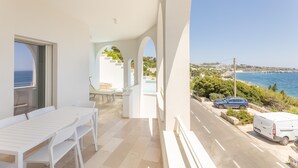 Een balkon met een witte tafel en stoelen, uitzicht op de zee en een weg met geparkeerde auto's.