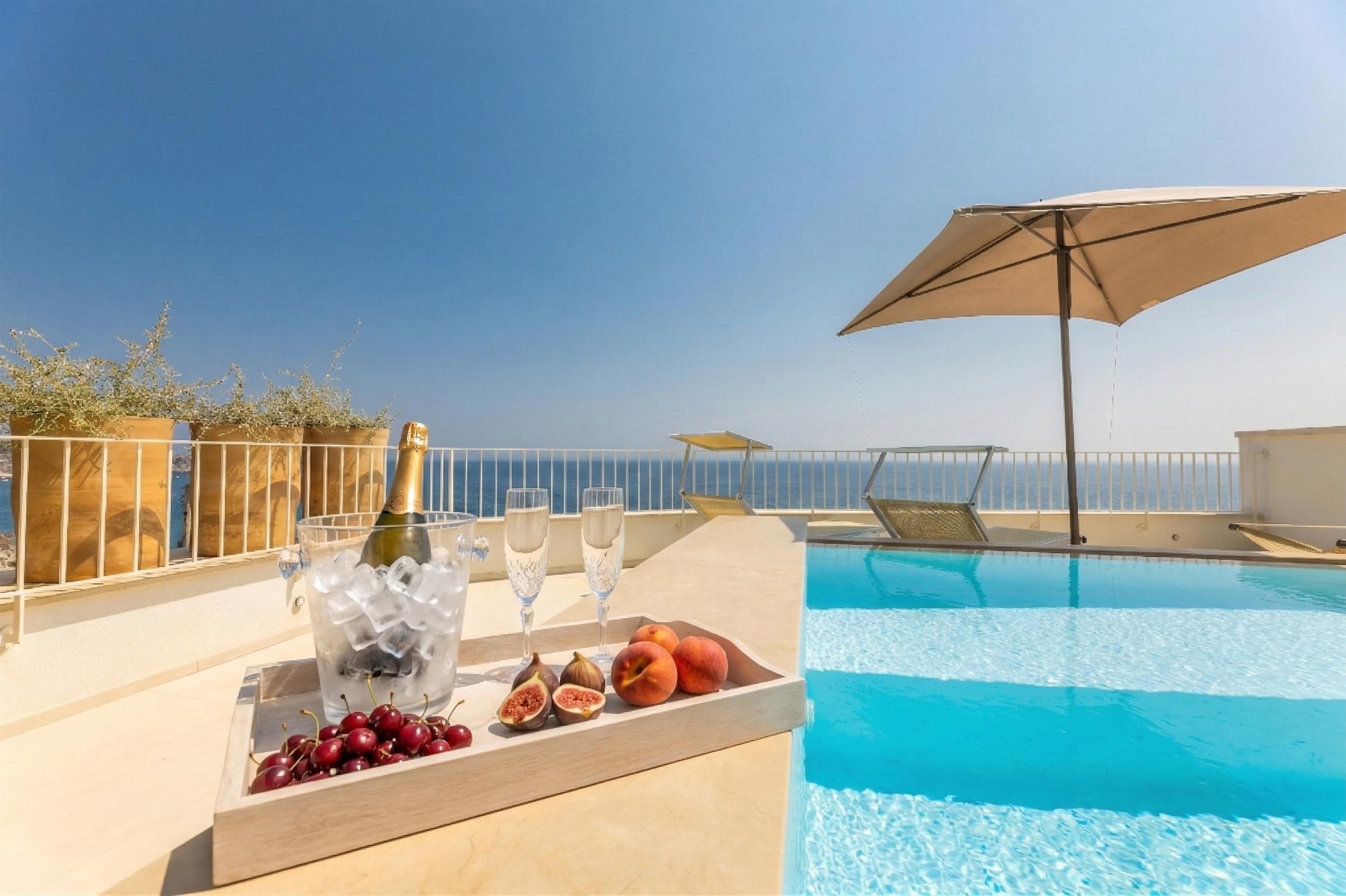 A poolside setting with a champagne bottle, glasses, and fruits on a tray, overlooking a clear blue sky and sea.