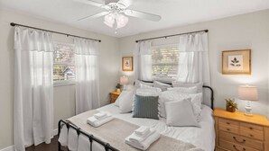 Un dormitorio ordenado con cama, mesitas de noche, ventilador de techo y cortinas.