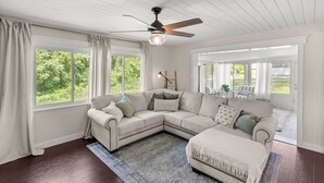 Amplia sala de estar con sofá modular, ventilador de techo y ventanas grandes que dan a vistas de vegetación.