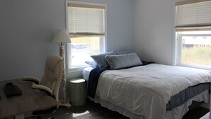 Ein Schlafzimmer mit einem Bett, einem Schreibtisch mit Stuhl, einer Lampe und zwei Fenstern mit Jalousien.